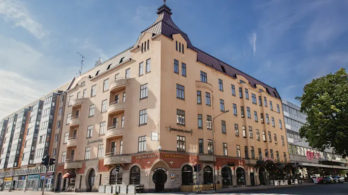 En beige fleretasjes bygning står på et gatehjørne med buede vinduer, balkonger og et bratt tak. Et tre og andre bygninger omgir det. Teksten lyder "Fennia Salonki" og "Fennia Restaurant".