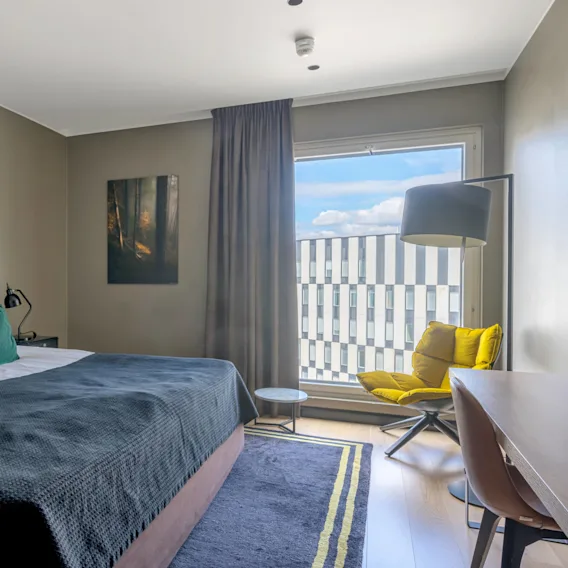 Siististi pedattu sänky vihreillä tyynyillä modernissa hotellihuoneessa. Keltainen tuoli ja iso ikkuna avautuvat kuvioitujen rakennusten näkymään.