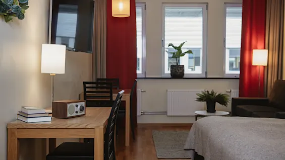 A desk with a lamp, books, and speaker sits next to a window; adjacent is a bed, sofa, and plants in a room with wood flooring and red curtains.