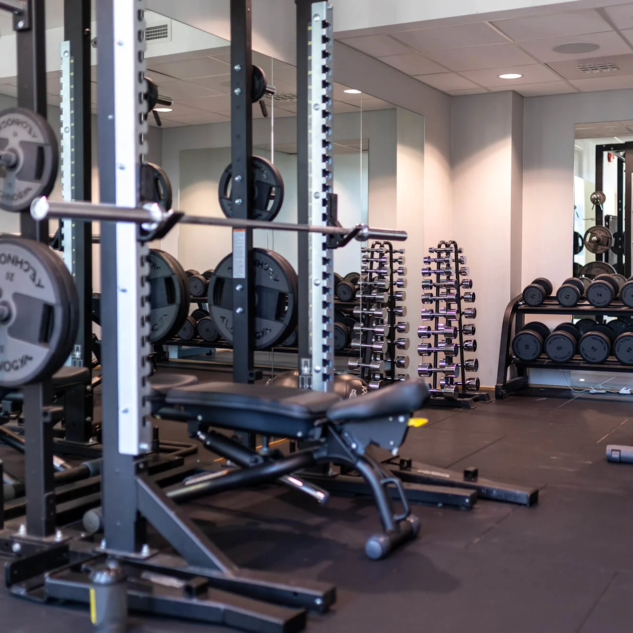 Ett gym med tyngdlyftningsutrustning, inklusive skivstänger på ett ställ, en bänk och hantlar arrangerade på ställ. Speglar reflekterar utrustningen och skapar en öppen och ljus atmosfär.