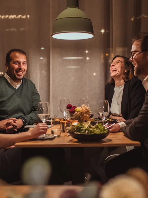 Fire venner som ler og prater rundt et middagsbord i et koselig og varmt opplyst restaurantlokale.