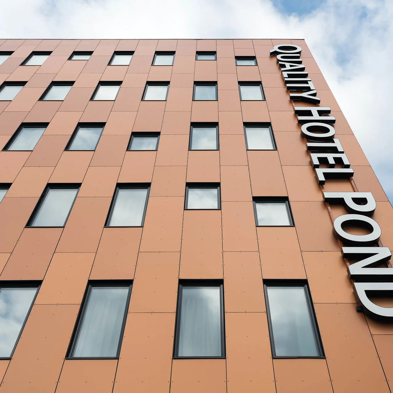 A tall building with orange panels and multiple windows displays the text "QUALITY HOTEL POND" on its facade under a cloudy sky.