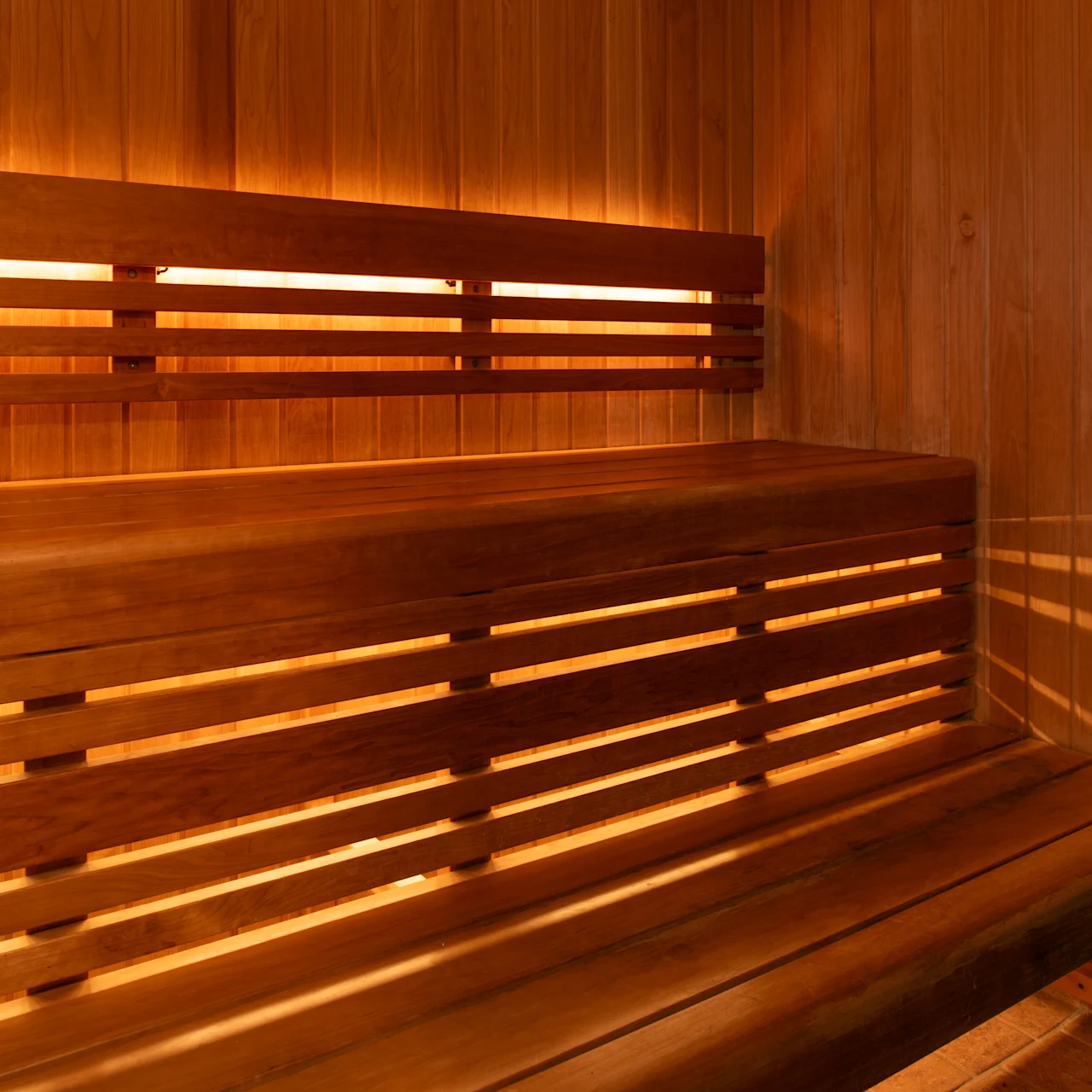 Træbeklædt sauna interiør med to niveauer af bænke langs væggene. Dæmpet belysning fremhæver den varme, hyggelige atmosfære. En metalovn med sten i hjørnet.