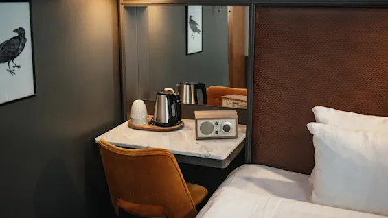 Et hyggeligt hotelværelse med et skrivebordsområde med en elkedel, kopper og en radio. En bog med titlen JACIE MILLS er synlig over spejlet.