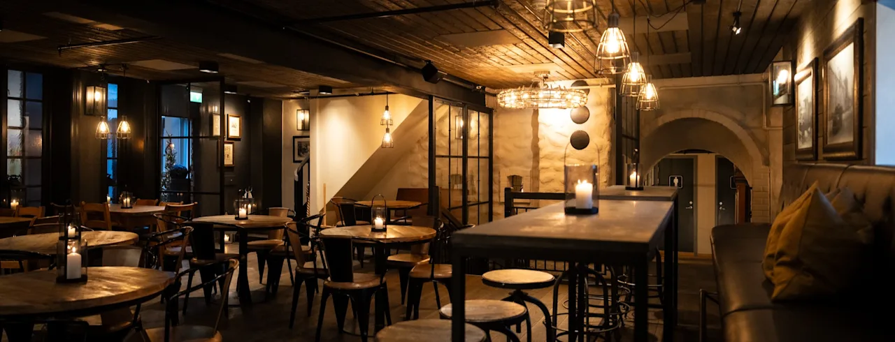 Et koselig, dempet opplyst restaurant eller bar med trebord, stoler og varm belysning fra stearinlys og taklamper.