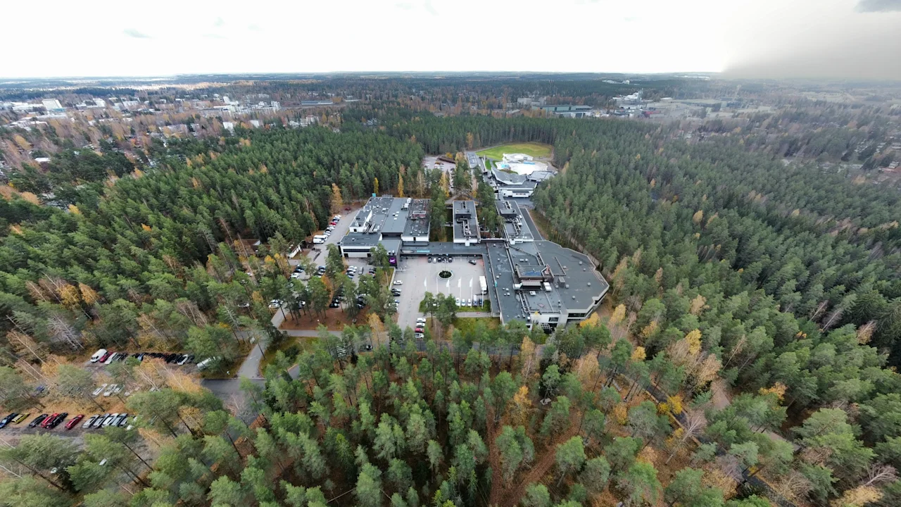 Suuri rakennuskompleksi on tiheän metsän ympäröimänä pilvisen taivaan alla, ja edustalla näkyy tie. Näkymä laajenee laajoihin metsämaisemiin.
