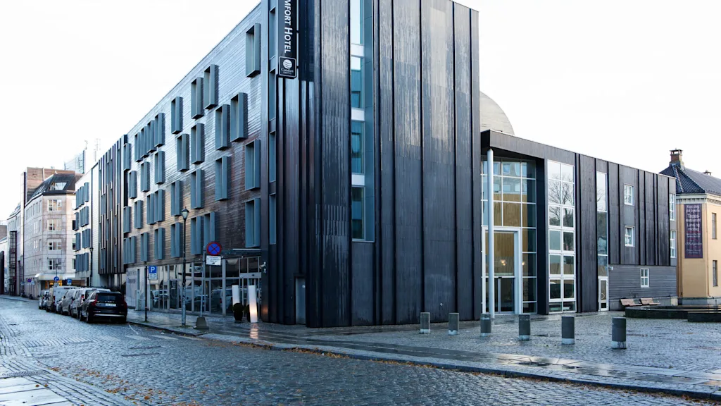 A modern building with a dark facade, displaying the sign "COMFORT HOTEL" vertically. It’s situated along a cobblestone street with parked cars and adjacent structures.