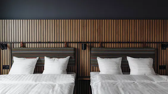 Two neatly made beds, topped with white pillows and linens, stand side by side against a vertical wooden slat headboard in a modern, minimalist room.