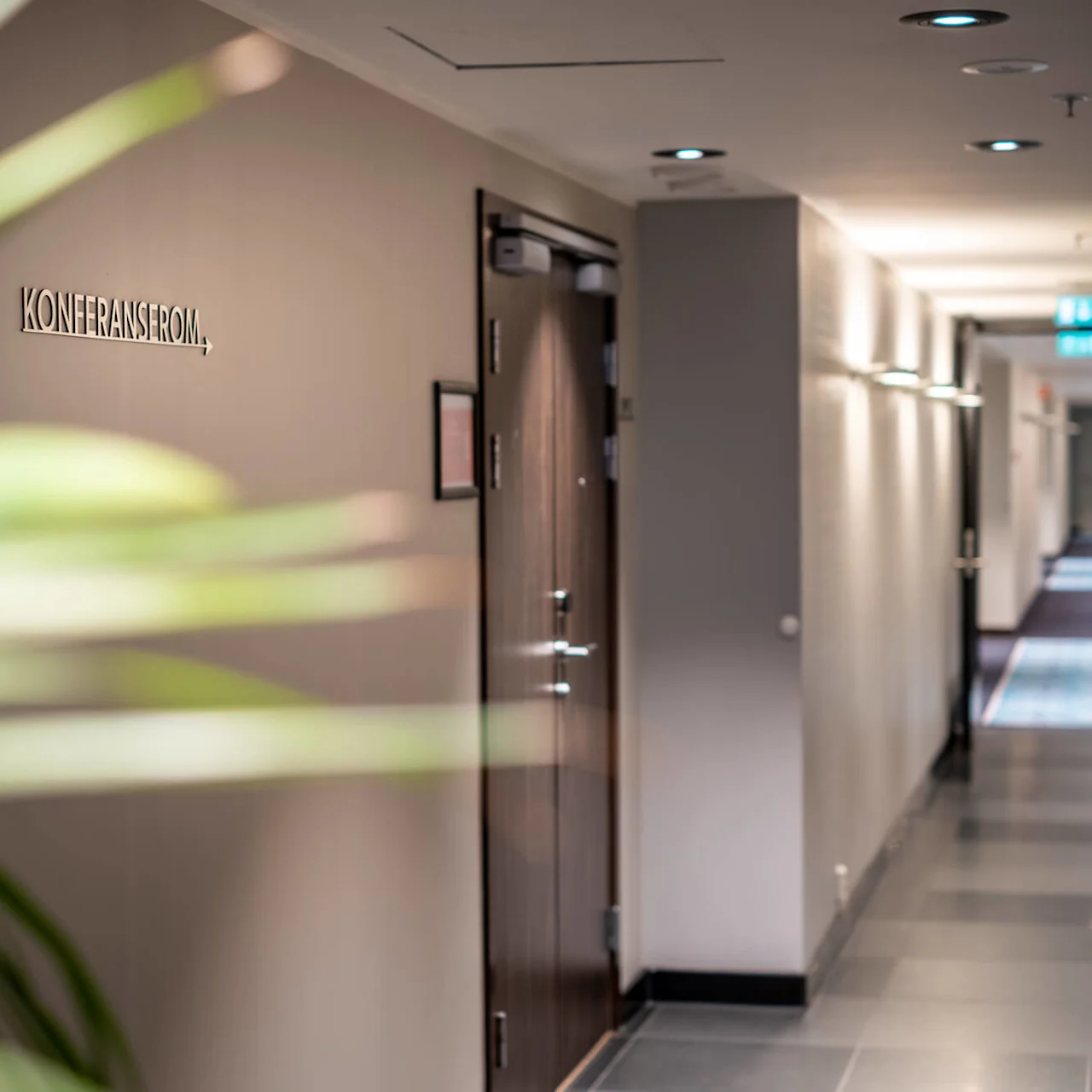 A hallway features a closed wooden door labeled "KONFERANSEROM," surrounded by beige walls and overhead lights, with a blurred green plant in the foreground.