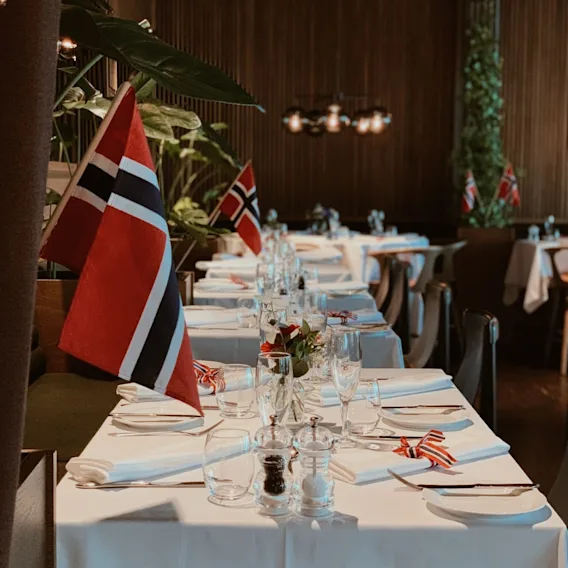 Et festdekket restaurantbord med hvite duker, vinglass, brettede servietter og små norske flagg.