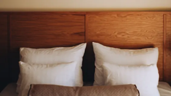 En hvit, rund papirlampe henger over en seng med myke hvite puter og en brun teksturert bolster, mot en trepanelbakgrunn.