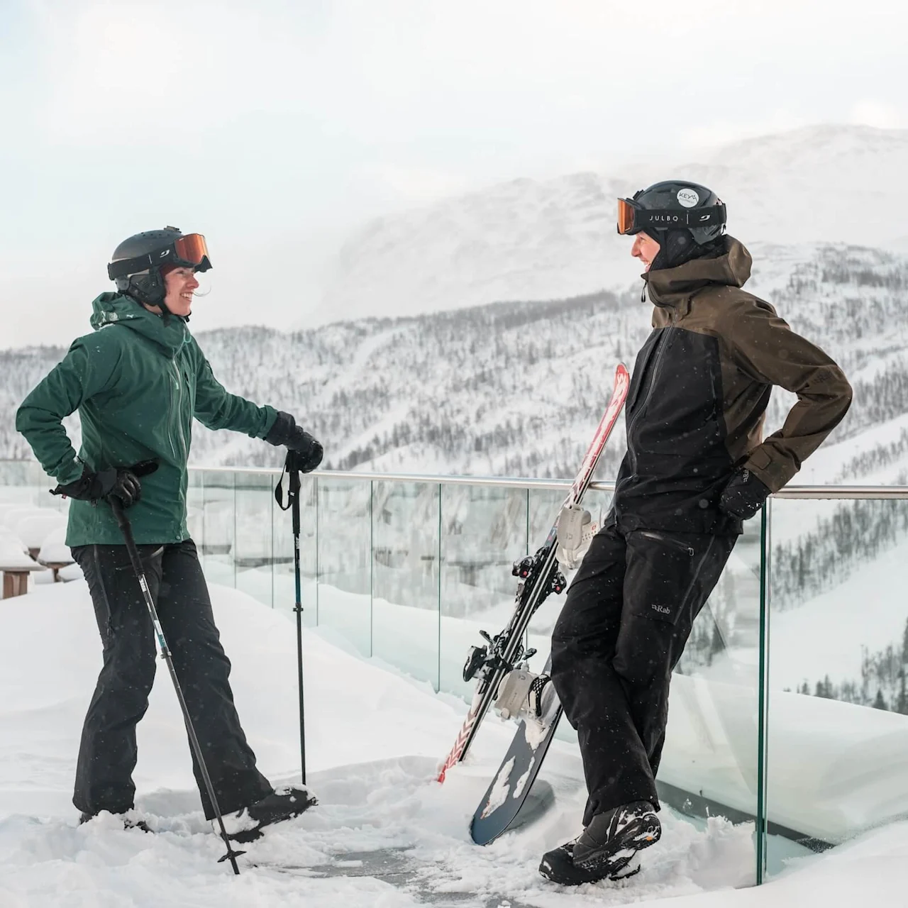 Two skiers standing on a snowy balcony, holding skis and poles, conversing. Snow-covered mountains and trees are visible in the background.