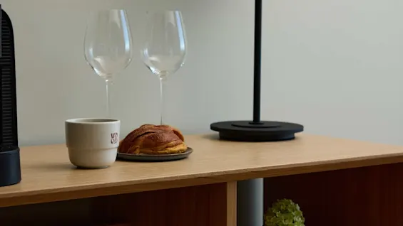 A modern table lamp with a layered shade sits on a wooden shelf beside two empty wine glasses, a coffee cup with "WOOD" text, and a pastry on a plate.