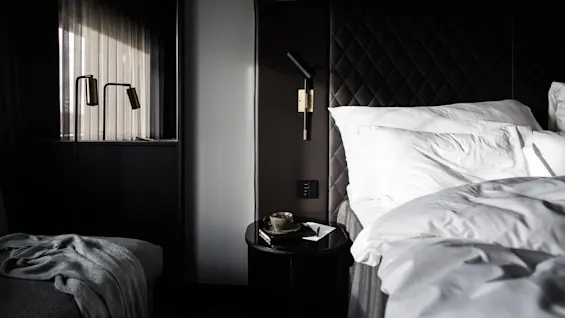Seng med hvite sengetøy står i et moderne, svakt opplyst rom. Et svart, quiltet sengegavl har en elegant lampe. I nærheten står et nattbord med en kopp og bok.