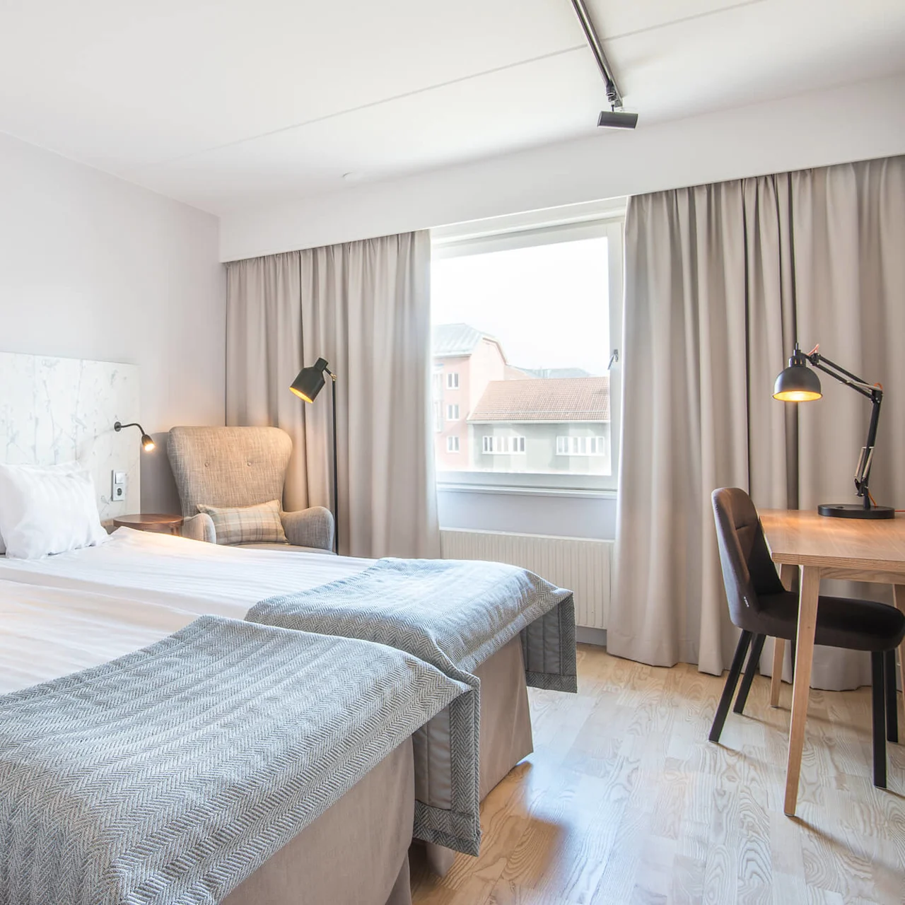 Två sängar med vita lakan och grå filtar är placerade i mitten av ett hotellrum, omgivna av sänglampor, en fåtölj och ett skrivbord. Ett fönster avslöjar byggnader utanför.