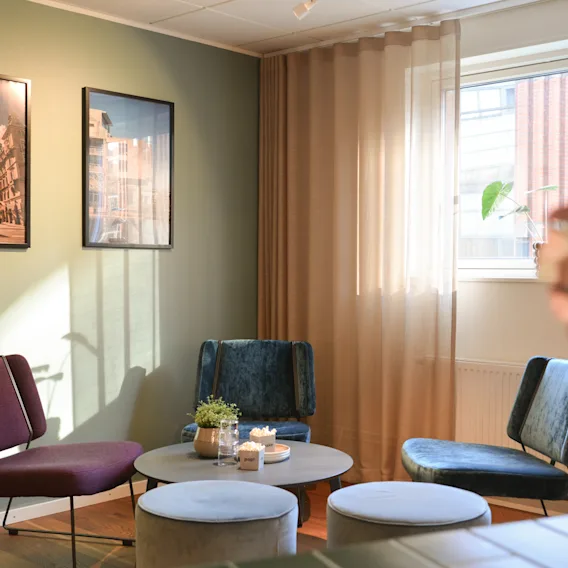 Purple and blue chairs encircle a round table with candles and a plant, set in a room with green walls and framed art. Sunlight filters through sheer curtains by the window.