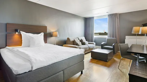 En seng med hvide sengelinned står i et moderne hotelværelse, ledsaget af en sofa, stole og et sofabord. Sollys filtrerer gennem et vindue med gardiner, som oplyser trægulvet.