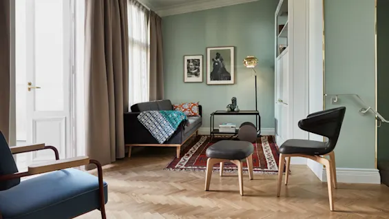 En svart lenestol står i en pastellfarget stue, dekorert med kunstverk, ved siden av en sofa, hyller og et lyst teppe. Lys strømmer gjennom åpne gardiner og en dør.