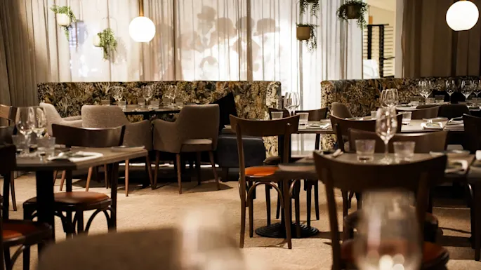 Bord og stoler er elegant arrangert i en svakt opplyst restaurant, omringet av hengende planter og dekorativ belysning, som skaper en koselig og intim atmosfære.