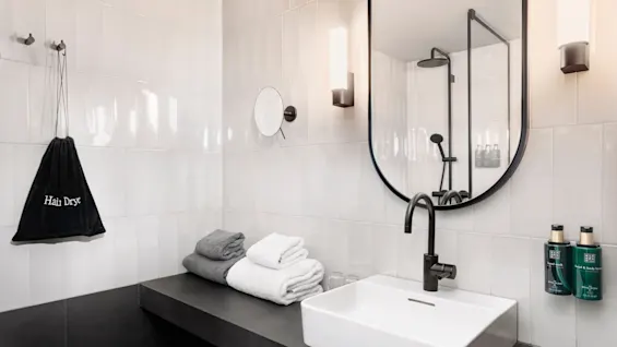 Handfat med svart kran står på en mörk bänk; handdukar är vikta bredvid. "Hair Dryer"-påse hänger på väggen i ett kaklat badrum med en spegel.