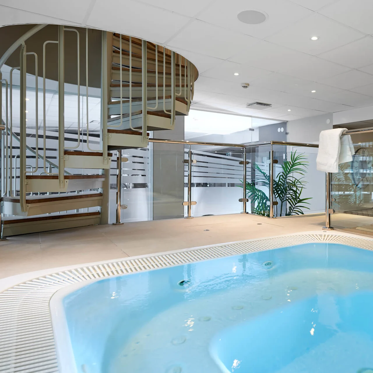 Whirlpool spa in a modern indoor setting with glass and metallic railings, spiral staircase, and plants.