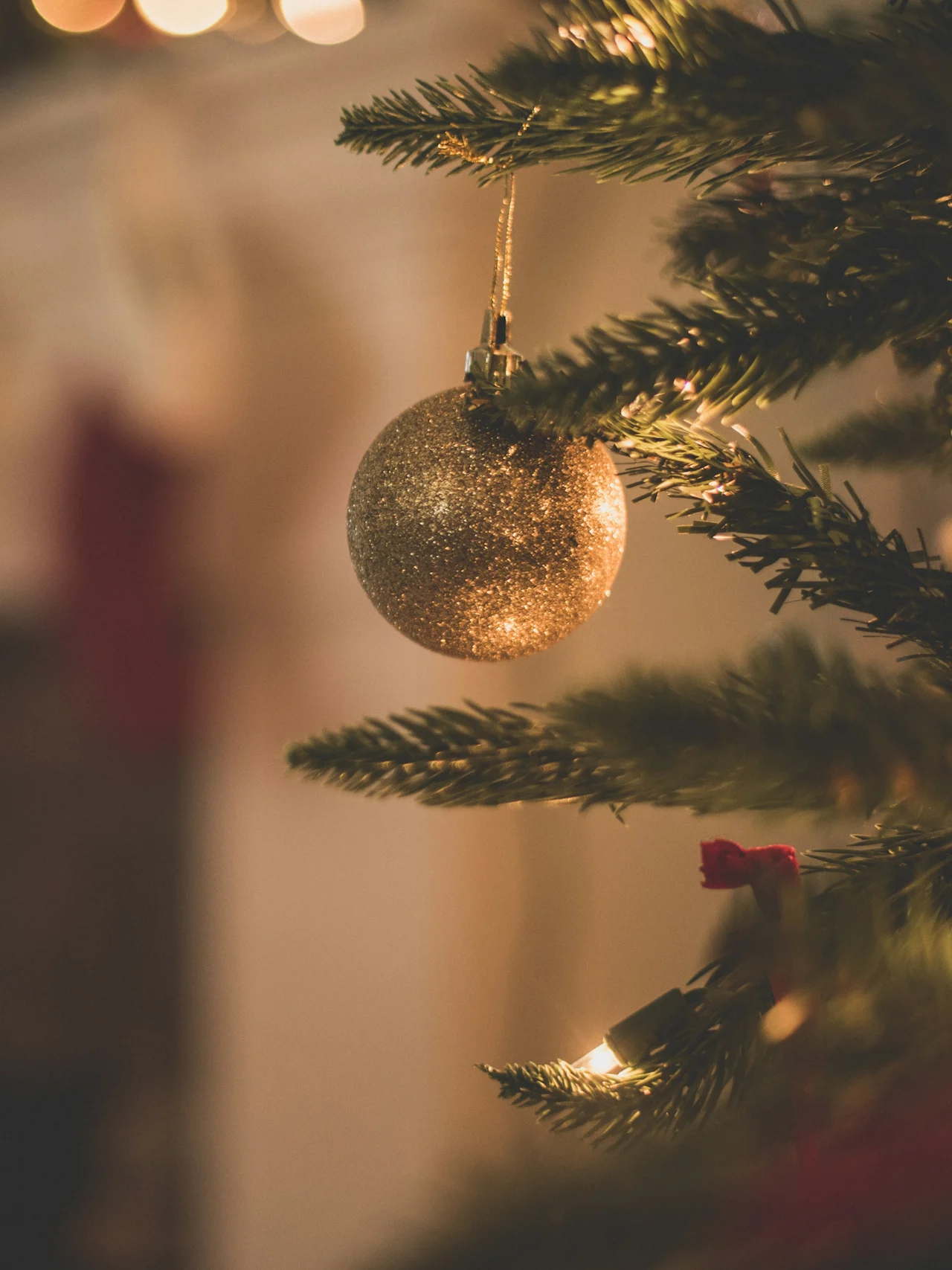 A golden bauble hangs from a Christmas tree branch, softly lit by string lights. In the background, Christmas stockings hang above a fireplace, creating a warm, festive atmosphere.