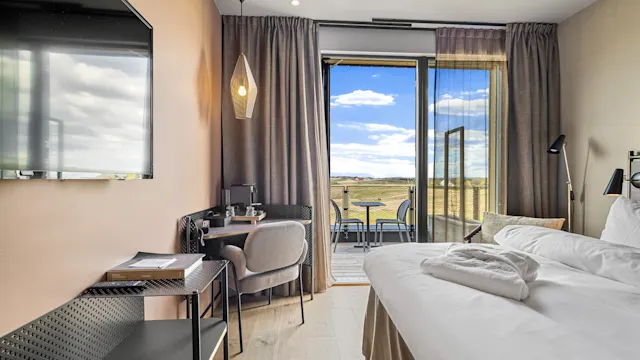 En pent senger i et moderne hotellrom med et skrivebord og en TV. Åpne skyvedører fører til en balkong med utsikt mot et rolig landskap under en blå himmel.