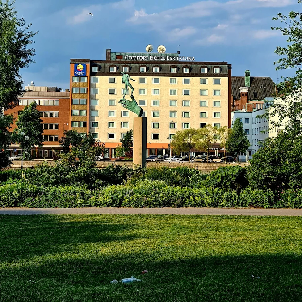Patsas seisoo jalustalla vehreyden keskellä, taustalla "Comfort Hotel Eskilstuna"-rakennus sinisen taivaan alla.