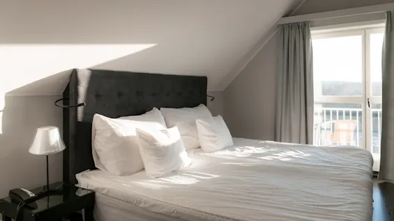 Et lyst og indbydende hotelværelse med en stor seng, hvidt sengetøj og sollys, der strømmer ind gennem en altandør.