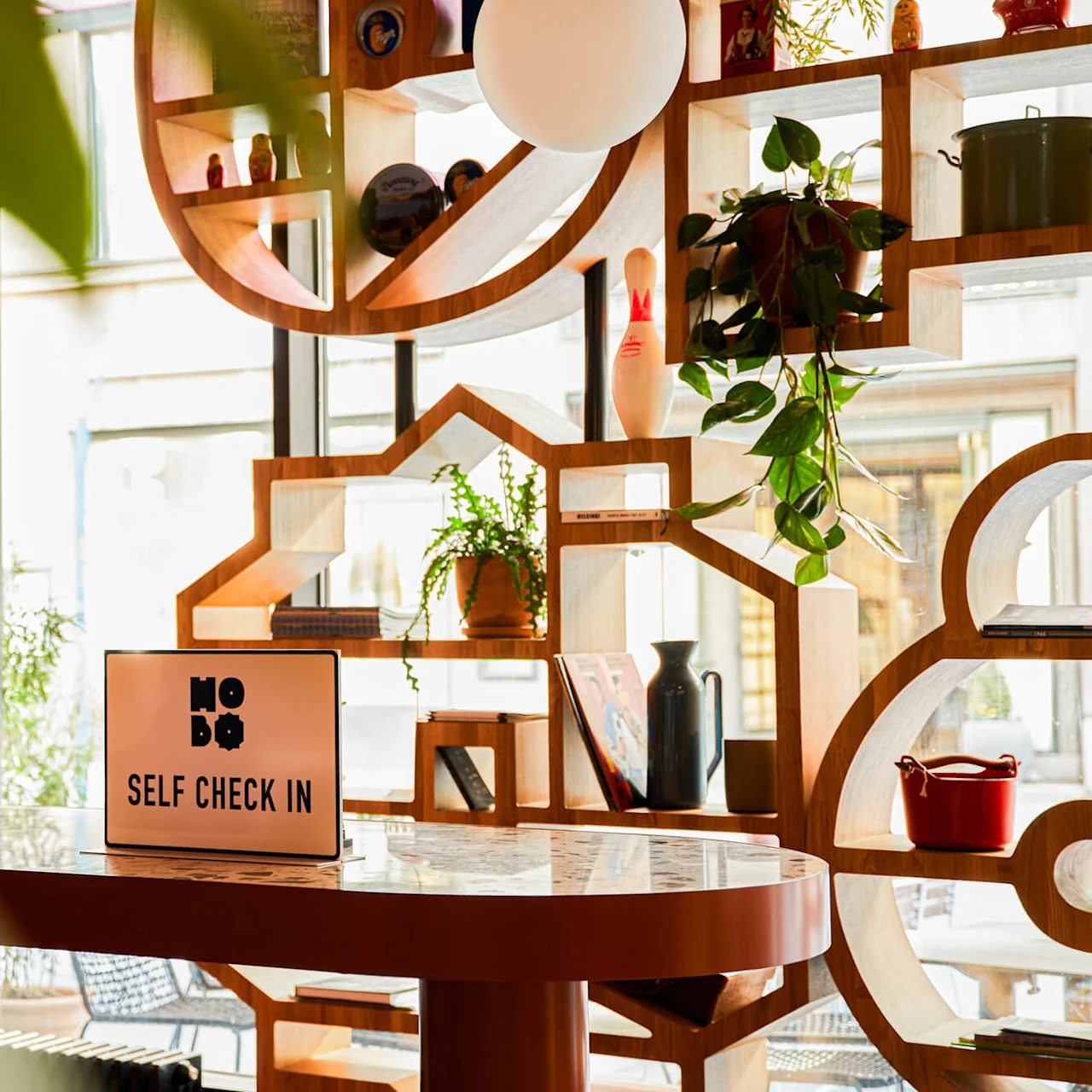 Et skilt på et bord lyder "HOBO SELF CHECK IN" i et moderne interiør med geometriske hylder, planter og dekorative genstande.
