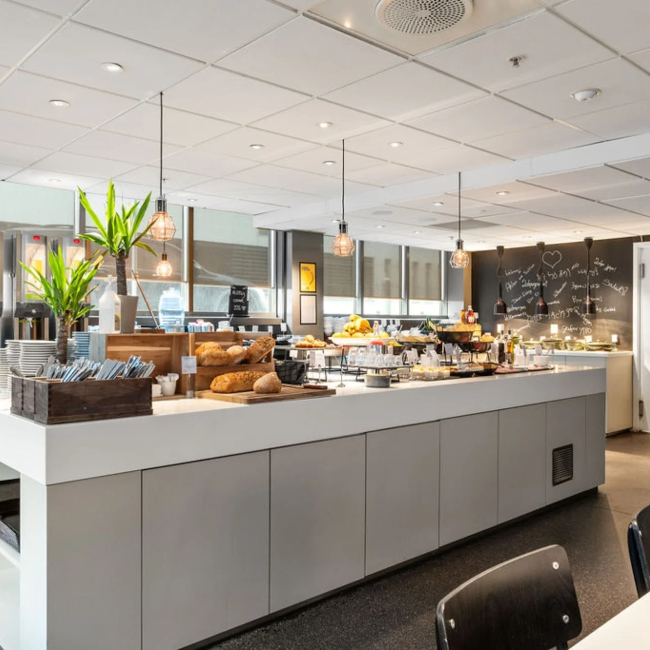 A self-serve buffet in a bright dining area features various foods and beverages on a large central island. Blackboards with unreadable text and drink dispensers are in the background.