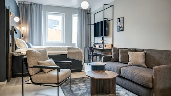 En modern hotellrum med en mysig säng och nattduksbord, en fåtölj och en soffa med ett soffbord. Ett skrivbord och en väggmonterad TV fyller hörnet.