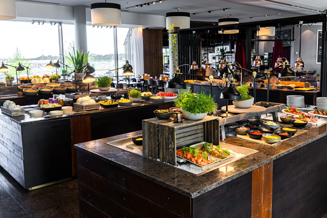 Et stort buffetbord med ulike retter inkludert salater, brød og garnityr. Det er omgitt av moderne innredning med store vinduer i en godt opplyst restaurantsetting.
