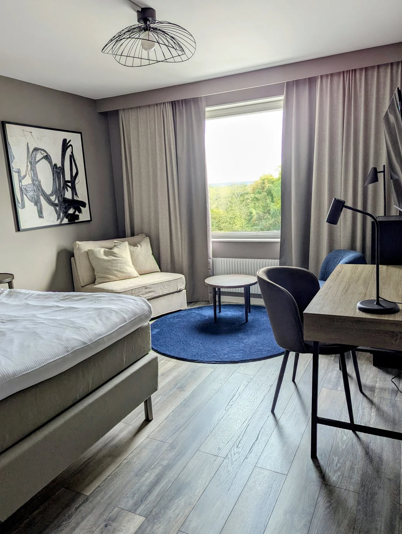 Ett modernt hotellrum med en bekväm säng, en mysig sittgrupp och ett skrivbord, med en fridfull utsikt över grönskande natur utanför fönstret.