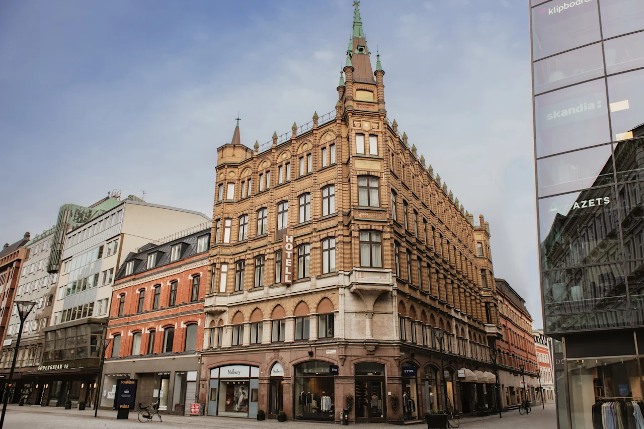 Home Hotel Baltzar. A grand, ornate brick building with a spire stands on a city street, with other buildings nearby. Text: HOTELL.