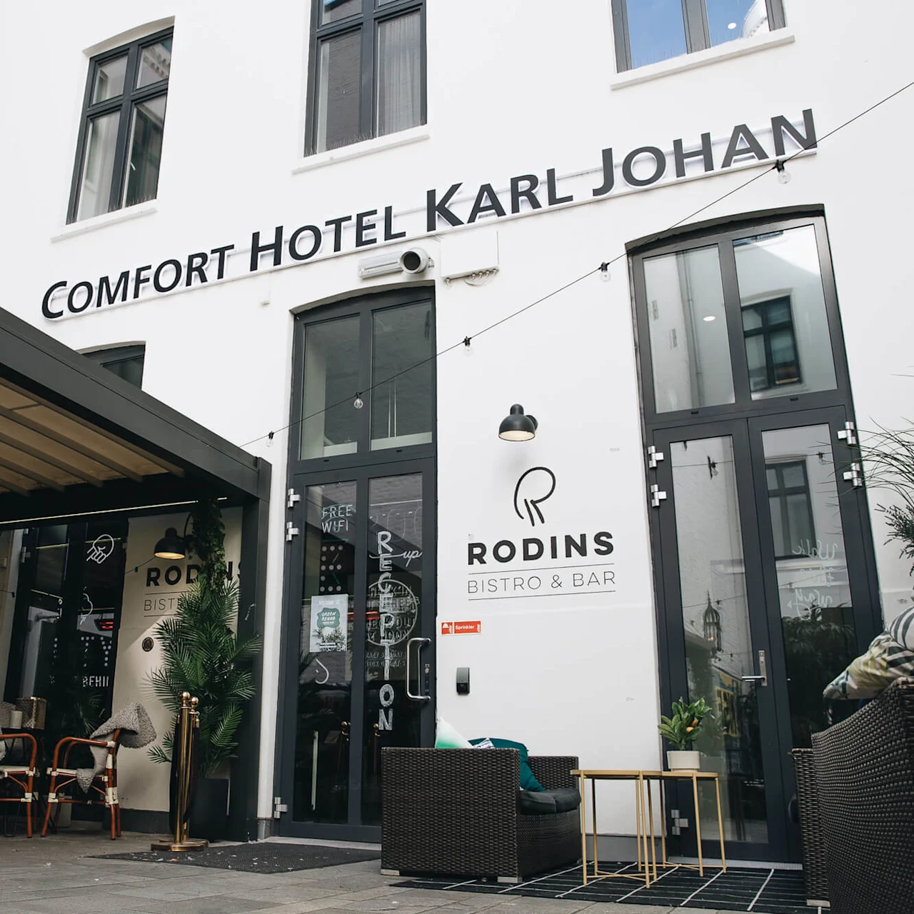 Et skilt lyder "Comfort Hotel Karl Johan" over dørene. En tavle med planter indikerer "Reception" med piler. Nærheten viser "Rodins Bistro & Bar". Bord og stoler er utenfor.