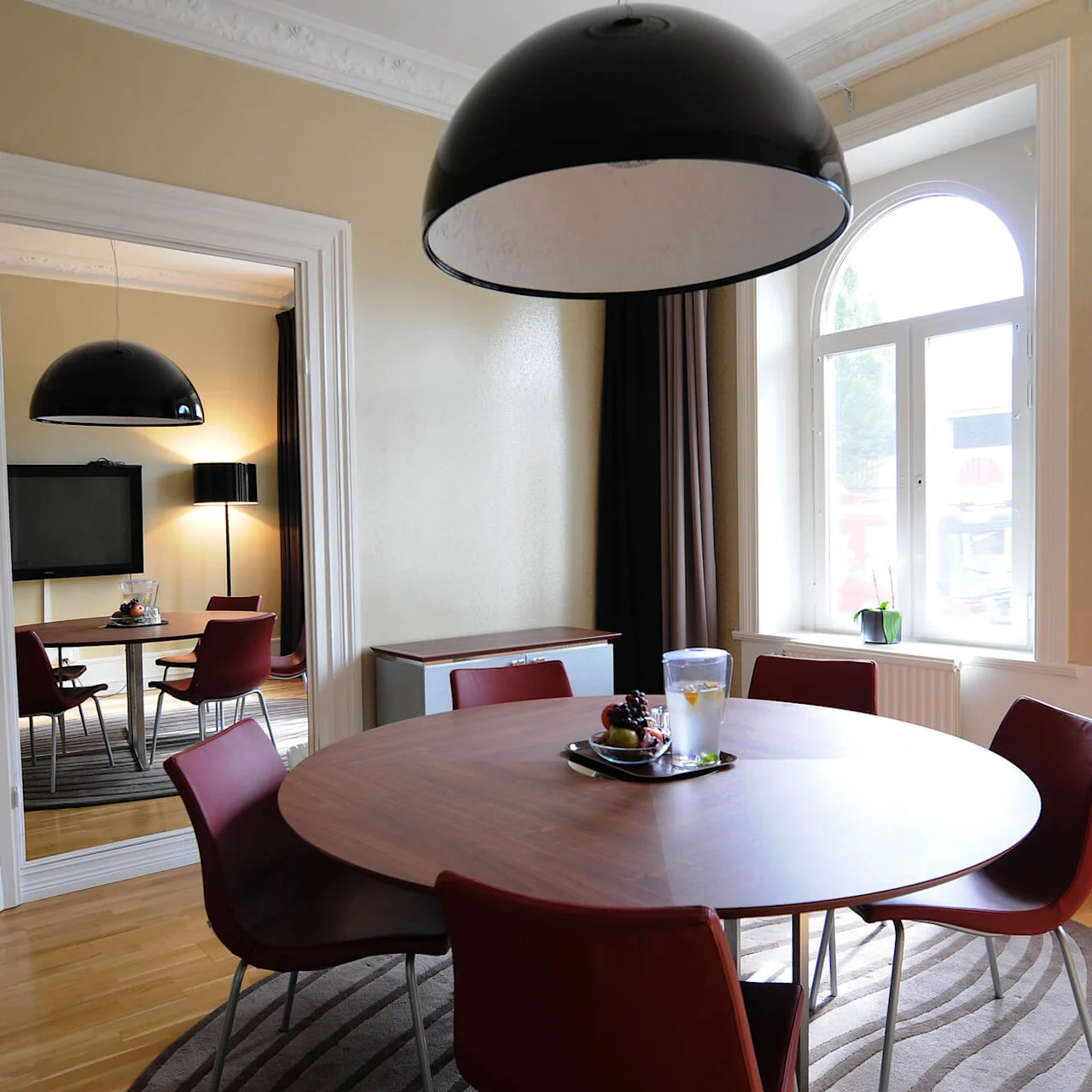 A round dining table with chairs, under a large pendant lamp, in a room with arching windows and ornate ceilings.