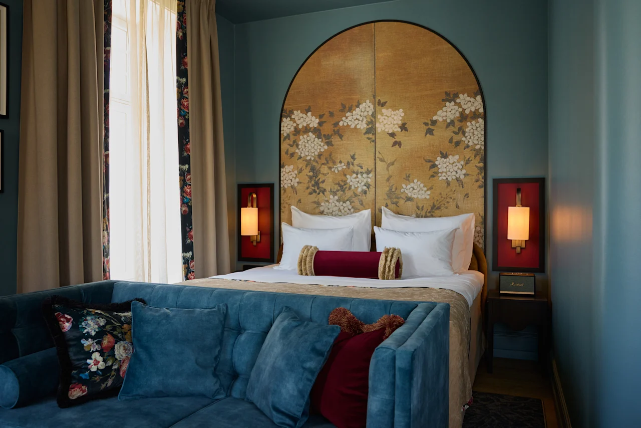 A bed with floral-patterned headboard sits in a cozy room. Two bedside lamps provide warm light. A blue sofa with decorative pillows faces the bed, creating a relaxing atmosphere.