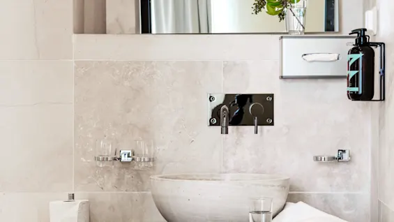 A stone sink rests on a dark marble counter with a glass of water and towel, set in a modern bathroom with light tile walls and mirror.