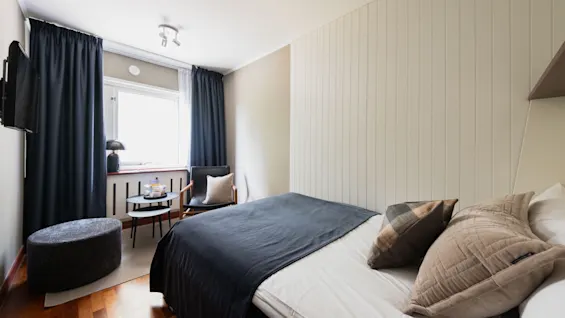 Et hyggeligt hotelværelse med en seng, et vindue med mørke gardiner, en behagelig stol og et lille bord.