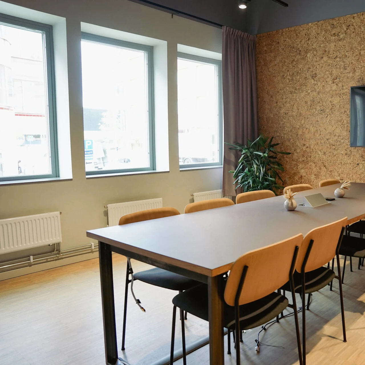 A modern conference room with a large table, chairs, and wall-mounted screen, illuminated by natural light from windows.