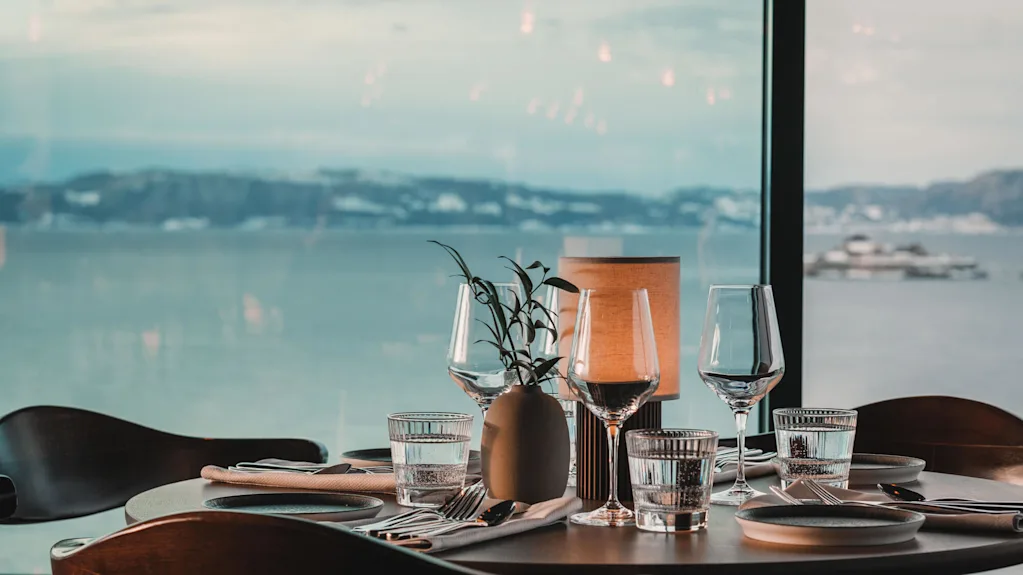 Ett bord är elegant dukat med glas, tallrikar och en liten växt, placerat vid ett stort fönster som visar en lugn havsutsikt med avlägsna kullar.
