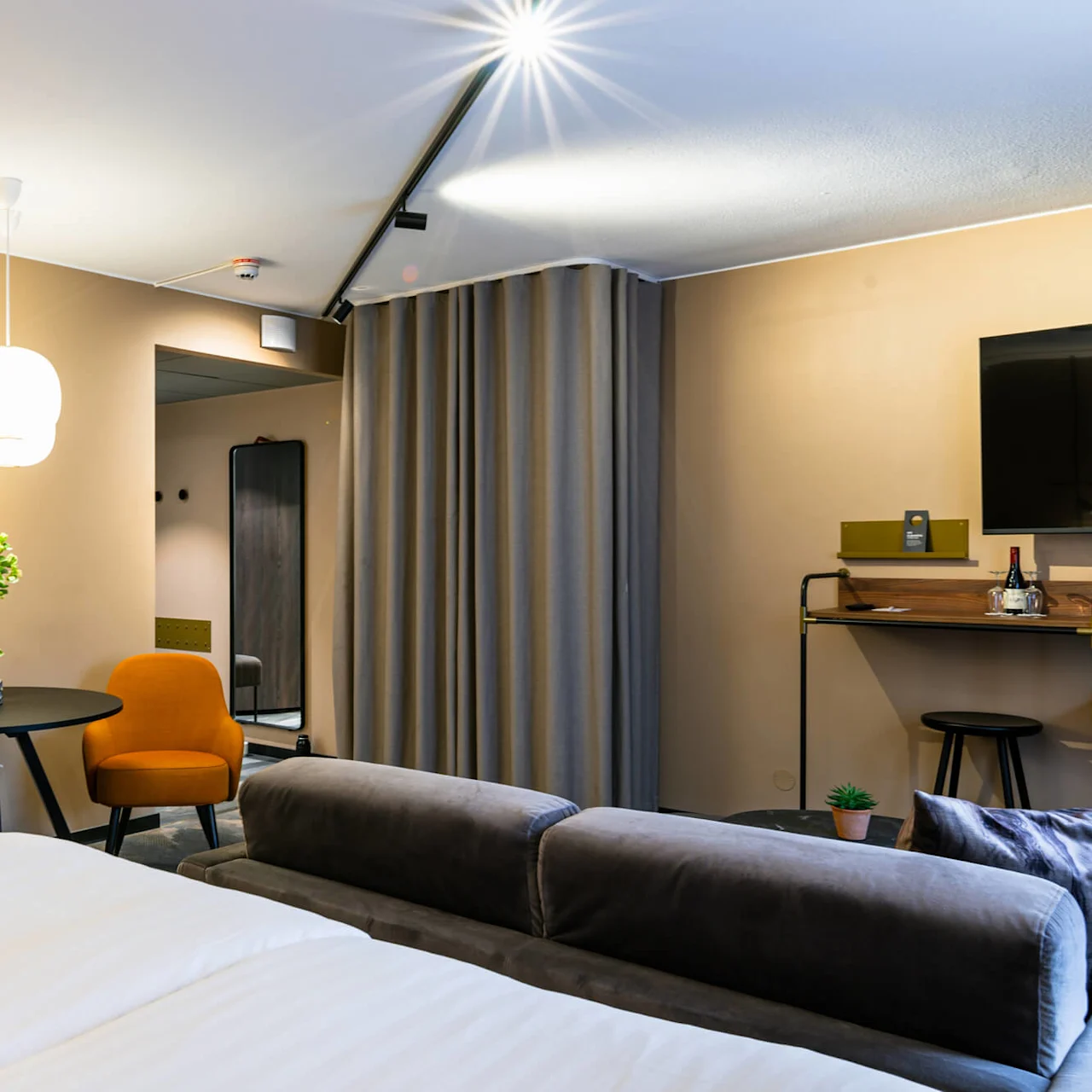 Et moderne hotelværelse med en blidt oplyst dobbeltseng, to orange stole ved et rundt bord med vaser og et vægmonteret TV over et skrivebord med vin og glas.