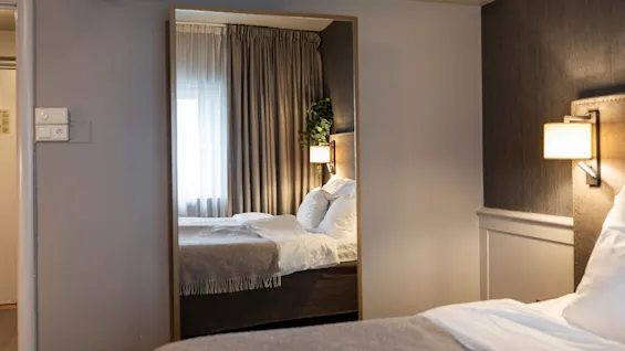 A comfortable hotel room at Home Hotel Grand Bodø with a large mirror reflecting the bed, curtains, and a wall lamp, creating a spacious feel.