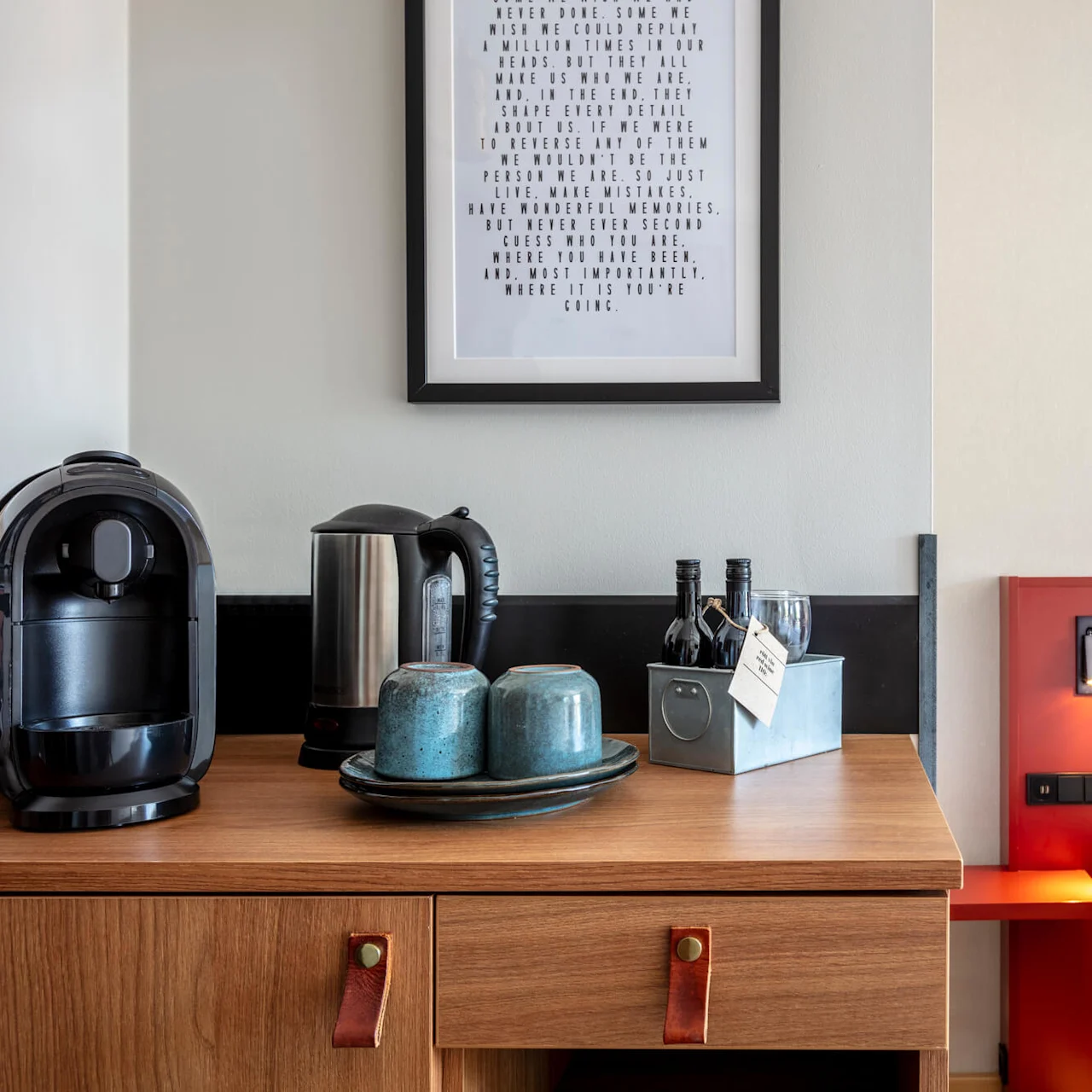 A hotel room's side table with a coffee maker and accessories against a wood-panel headboard.