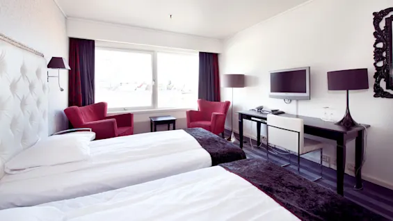 A well-lit hotel room with two single beds, red chairs, a desk, flat-screen TV, and a large window.