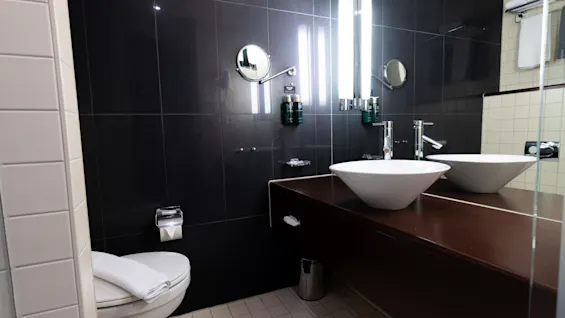 A modern bathroom features two white vessel sinks on a dark wooden counter, with illuminated mirrors above. A toilet with a folded towel lies nearby, surrounded by dark tiled walls.