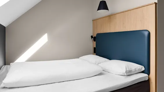 En säng med vita lakan står under ett lutande tak. En träpanel stödjer en mörkblå kudde. En svart pendellampa hänger i närheten.