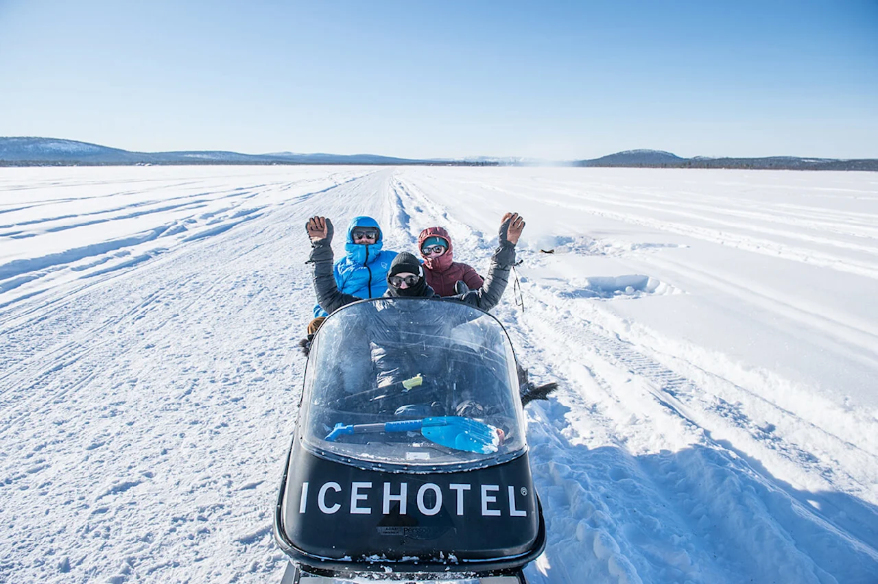 Snescooter mærket "ICEHOTEL" med tre personer vinkende, kørende gennem et snedækket landskab under klare blå himmel.