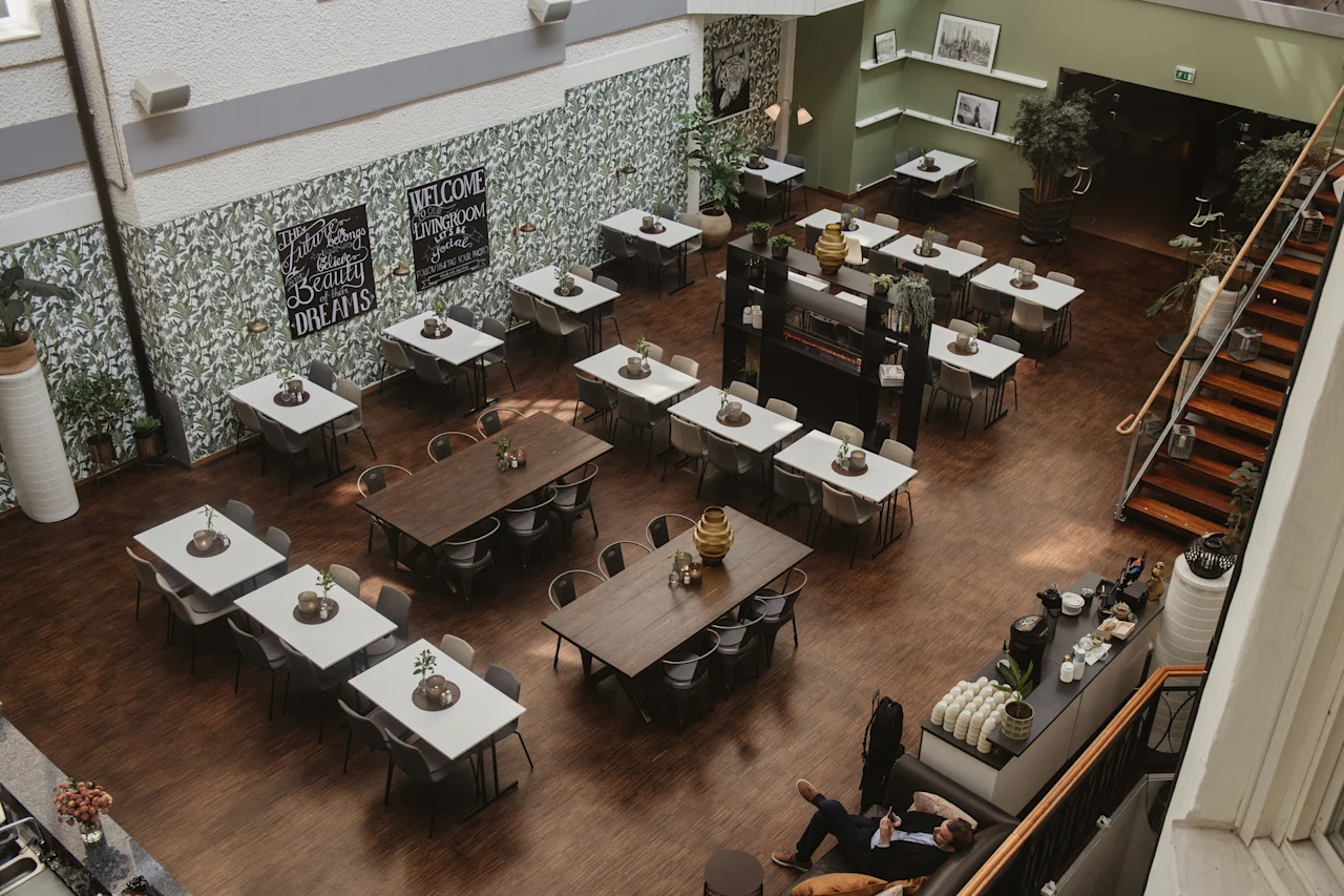 Borden är arrangerade med vaser ovanpå i en rymlig, väl upplyst matplats med grön och vit inredning. Trappor leder uppåt. Skyltar lyder: "Cafeteria, Talk About Your Dreams," "Welcome Lounge."